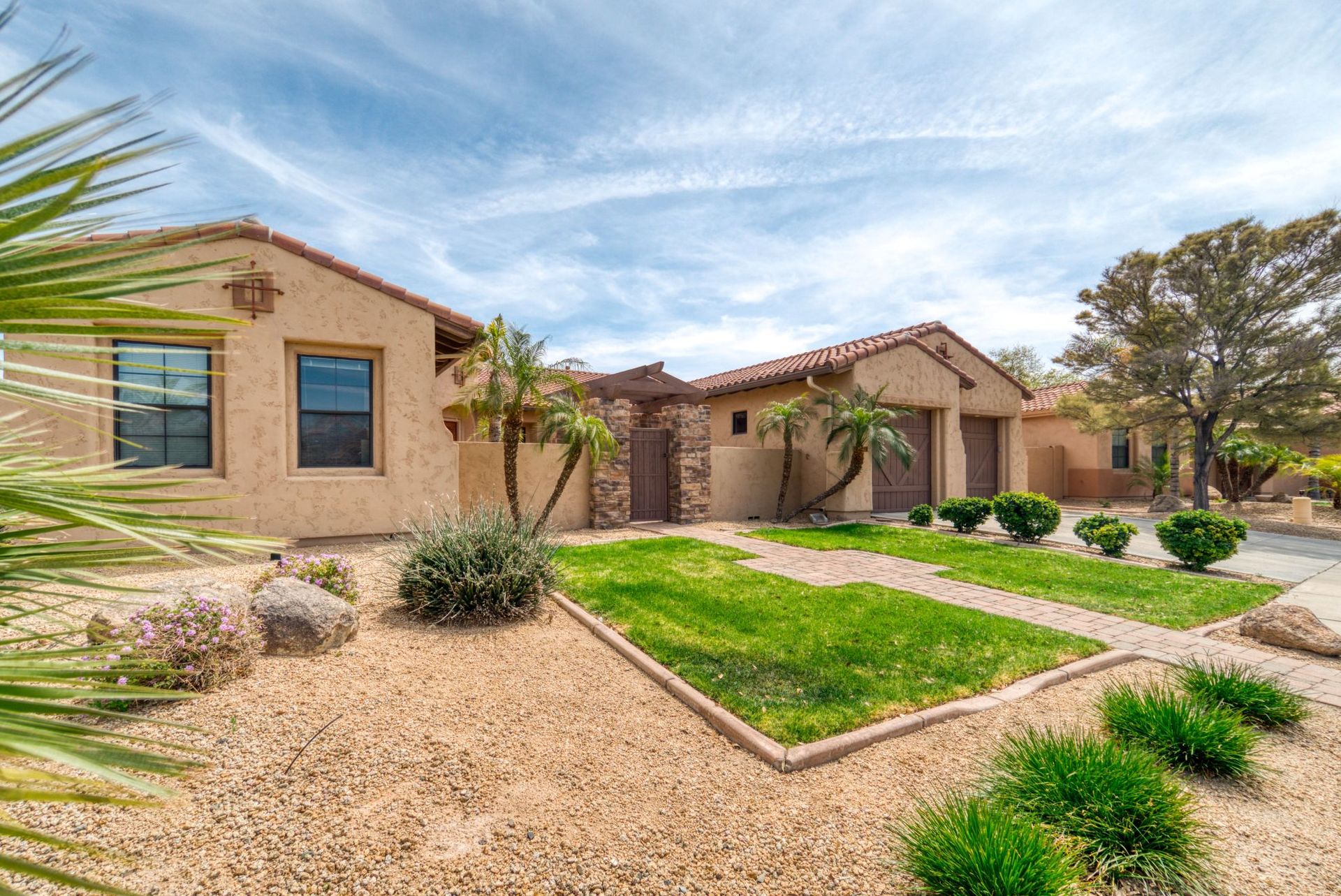 Desert-style stucco house with a landscaped front yard, gravel paths, green lawn, and palm trees