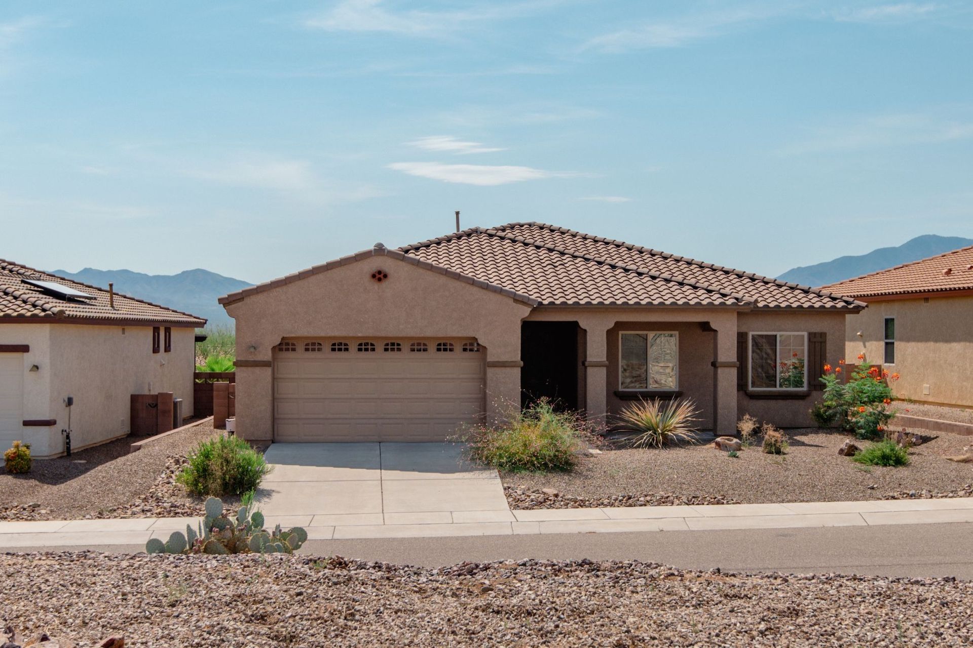 Single-story beige stucco house with tiled roof, garage, and desert landscaping under a clear sky