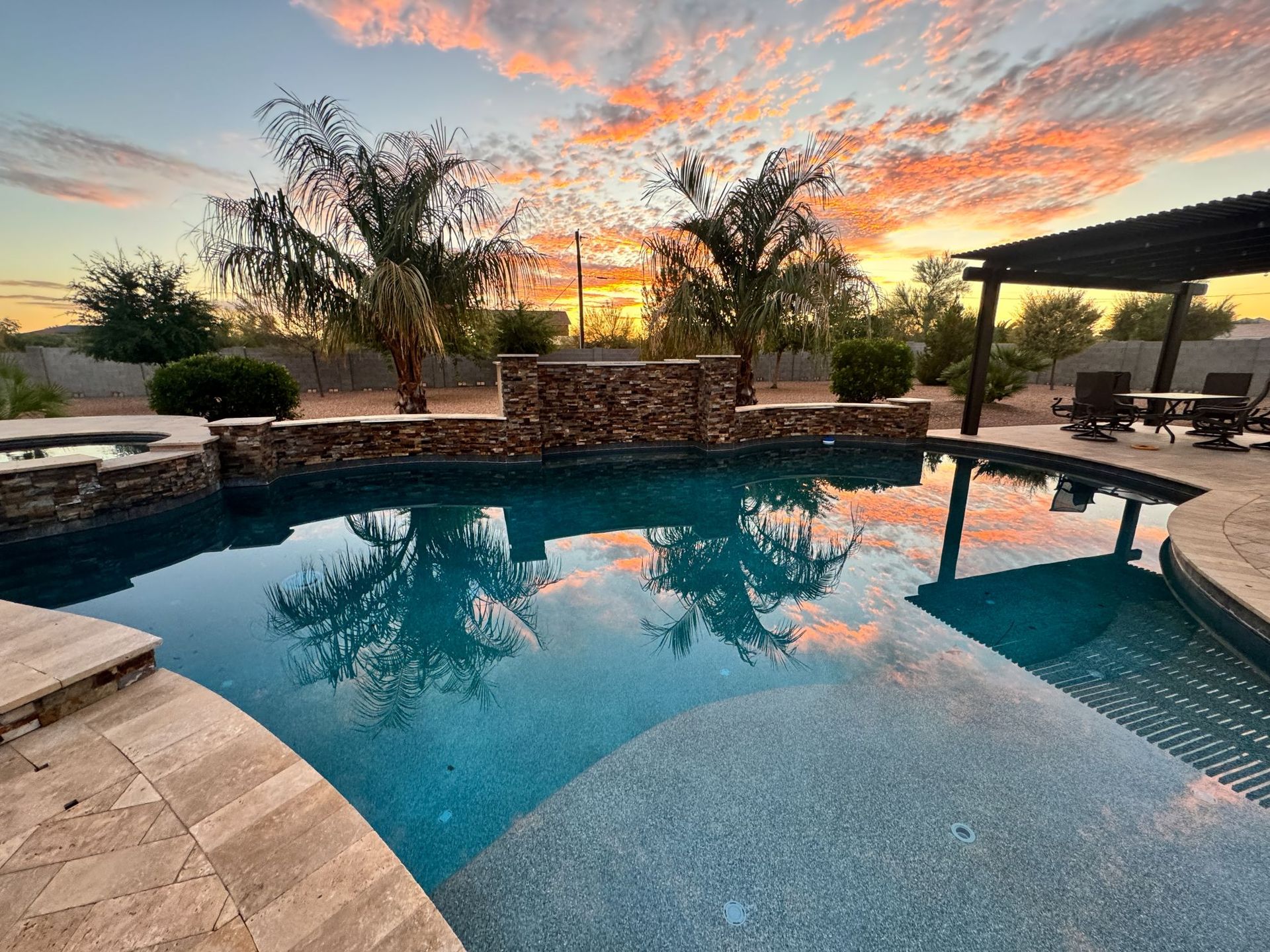 Backyard pool at sunset with stone patio, palm tree, and warm orange clouds reflected in the water