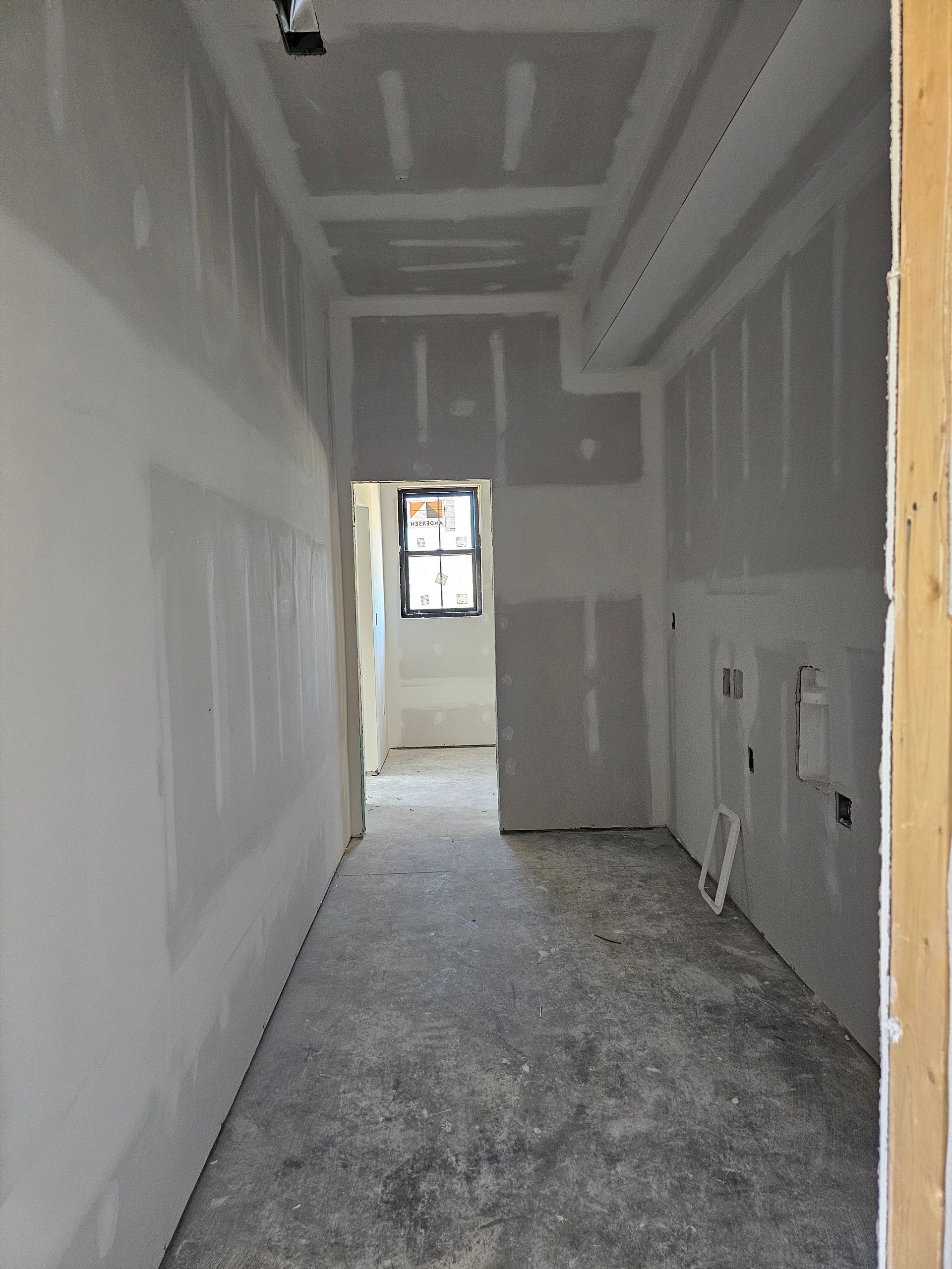 Interior construction of a hallway. Drywall walls and ceiling are in place. Concrete floor. Window visible at far end.