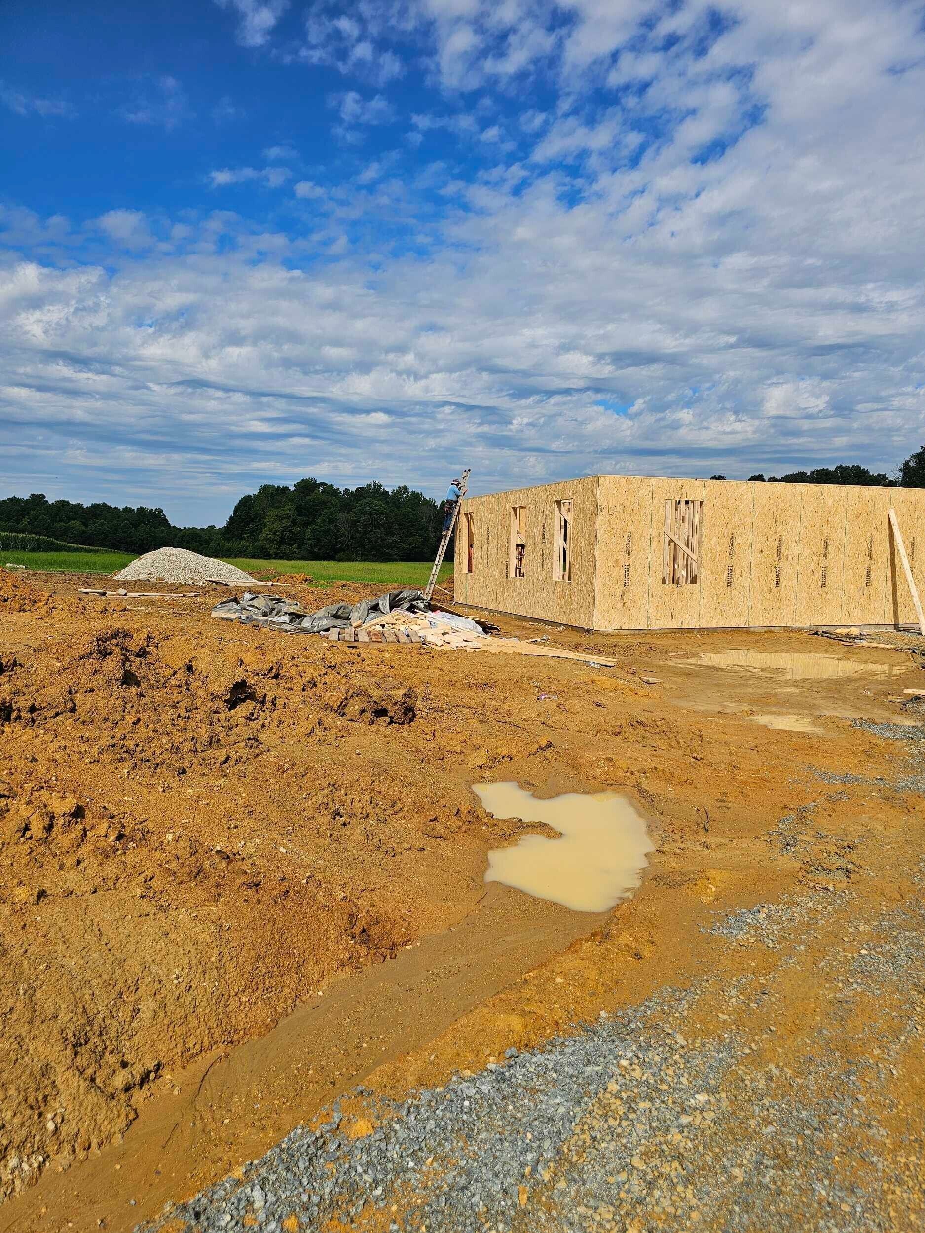 Dirt construction site with a muddy puddle, gravel road, and unfinished stone building under a cloudy sky
