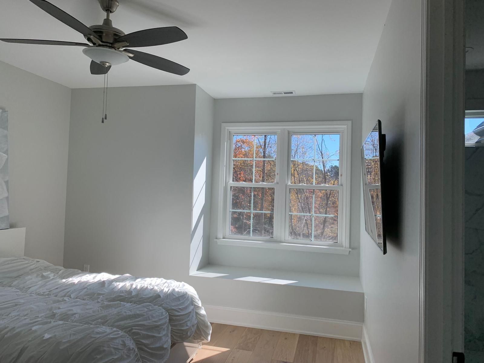 Bedroom with white bed, ceiling fan, and large window with sunlight and trees outside