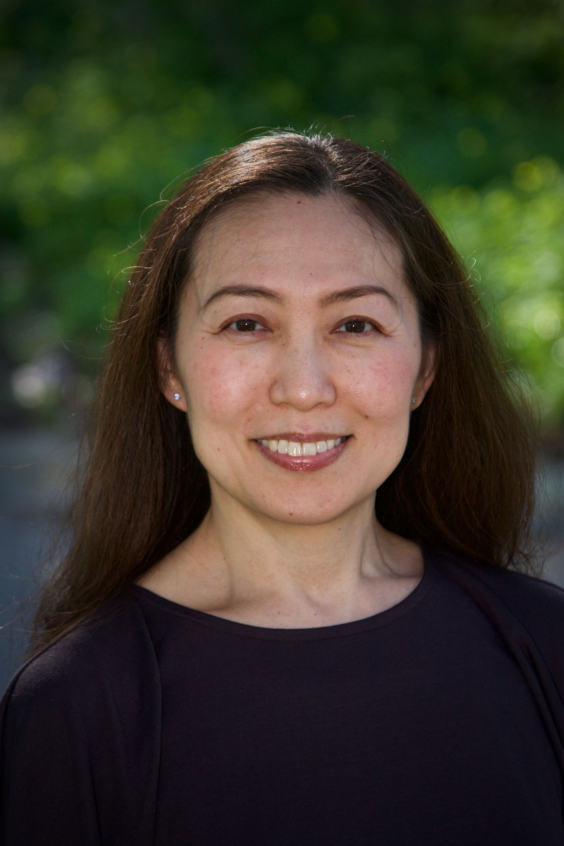 Smiling person with long dark hair in a black top, standing outdoors against a blurred green background