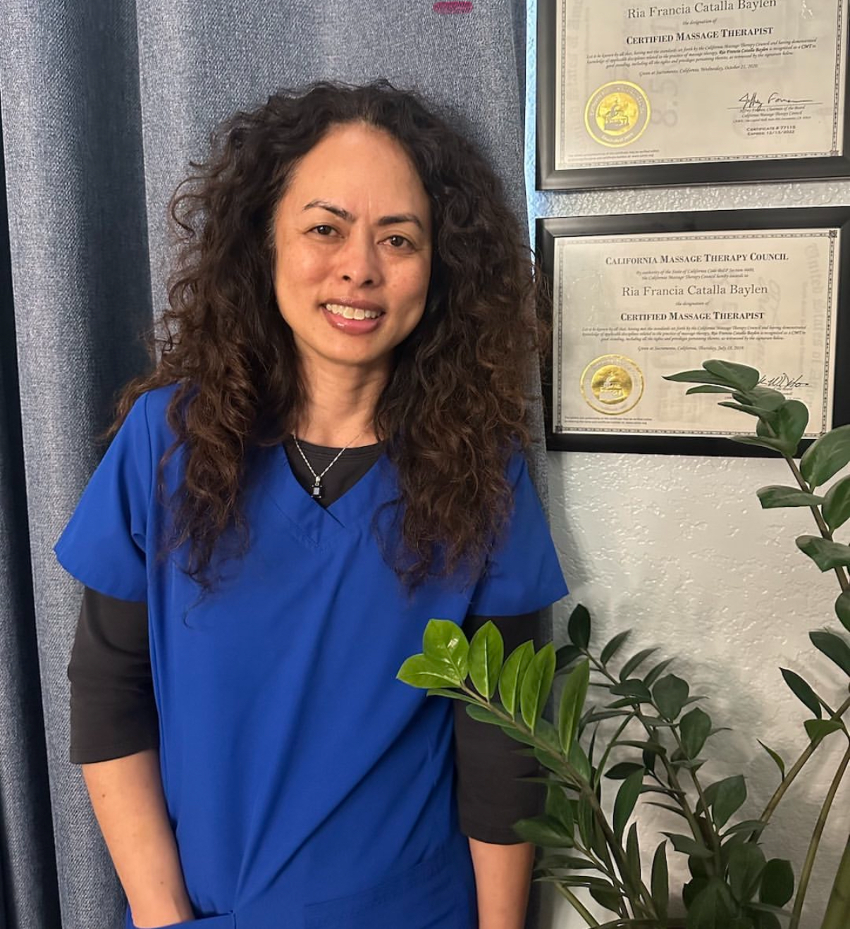 Woman in blue scrubs smiles near diplomas and a plant.