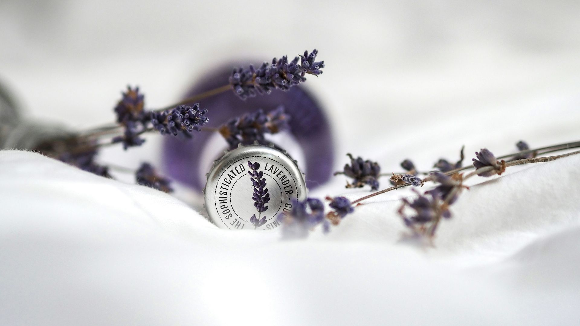 Purple lavender sprigs around a silver jar on soft white fabric, with a blurred purple sphere in the background