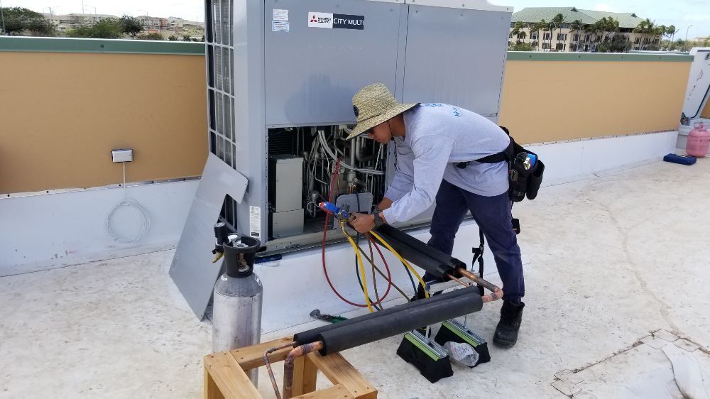 A man in a hat is working on an air conditioner on a roof.