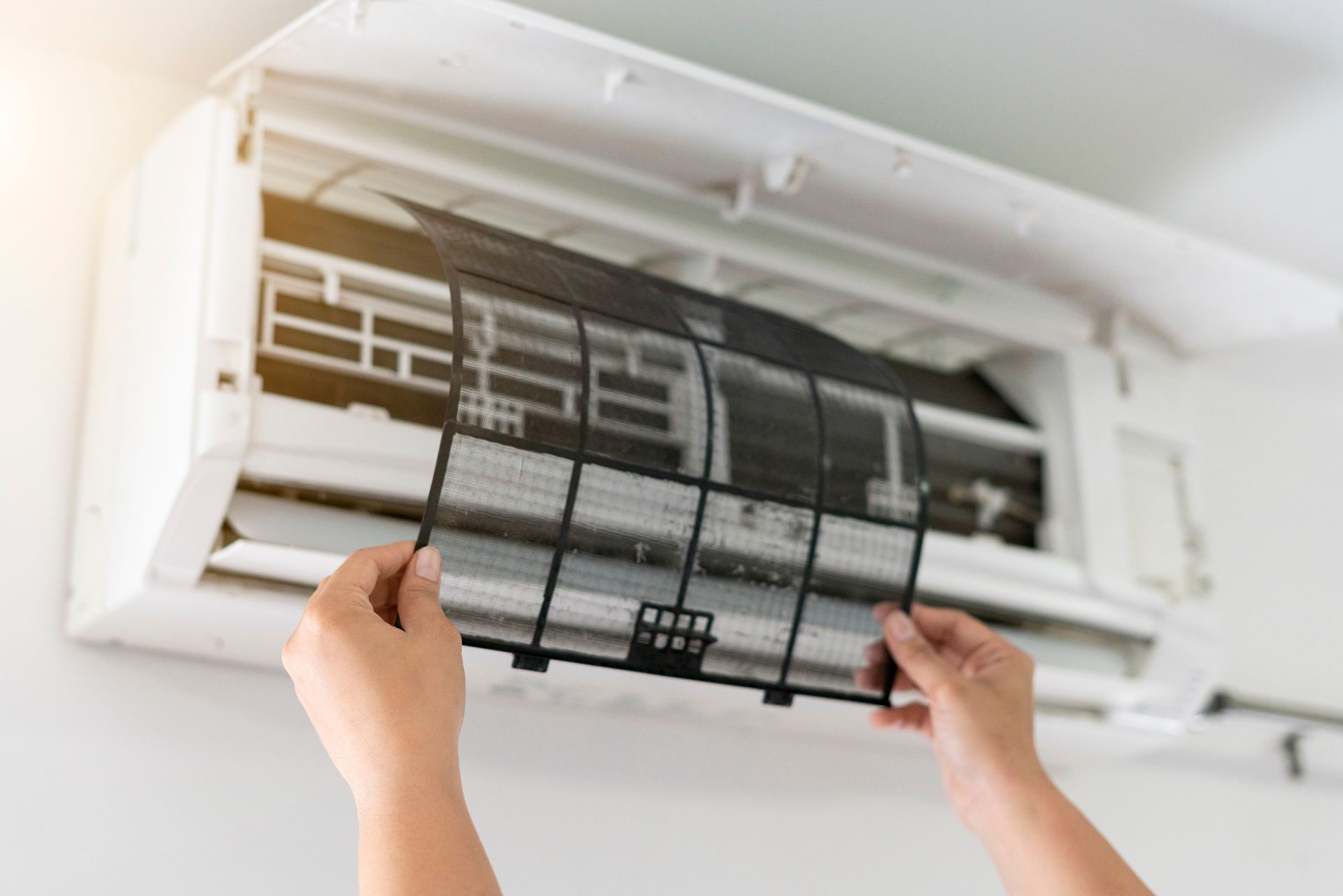 Hands removing an air conditioning filter from a white wall-mounted unit.