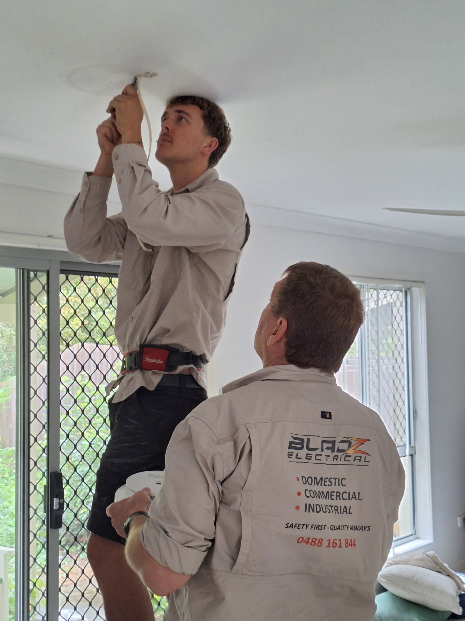 Two men installing a ceiling fixture indoors, one standing on a ladder while the other steadies him.