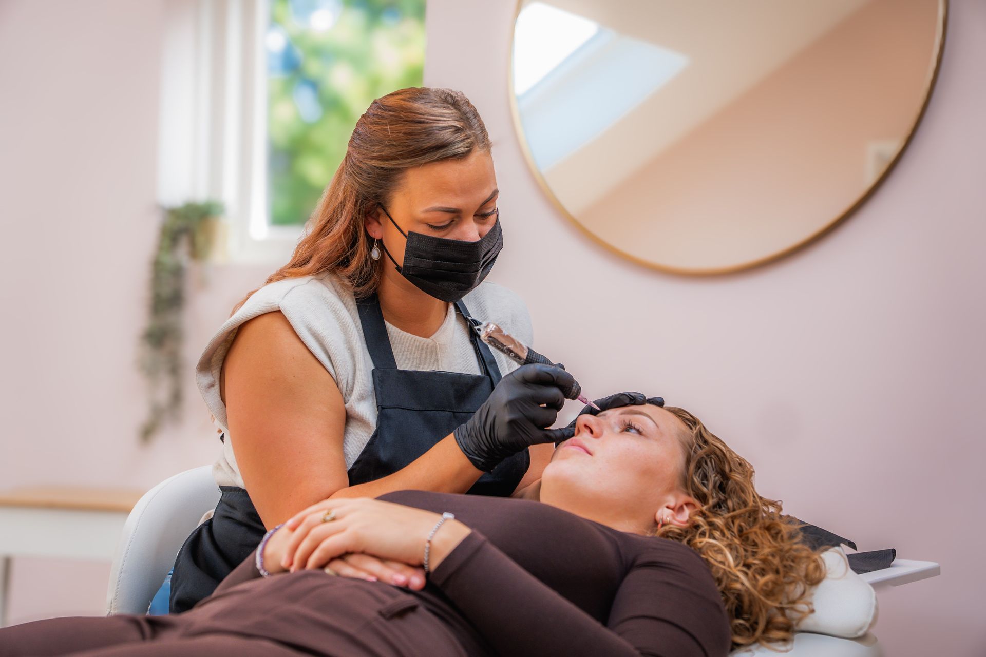 Een persoon met een masker werkt aan de wenkbrauwen van een klant; ze bevinden zich in een salon met een spiegel.