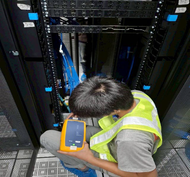 Technician in a reflective vest inspecting server racks in a data center with a handheld tester.