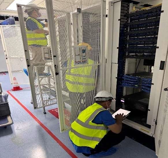 Workers inspecting equipment in a server room, one crouching by an open rack with a clipboard.