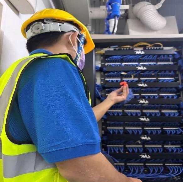 Technician in hard hat and mask inspects blue network cables in a server rack.