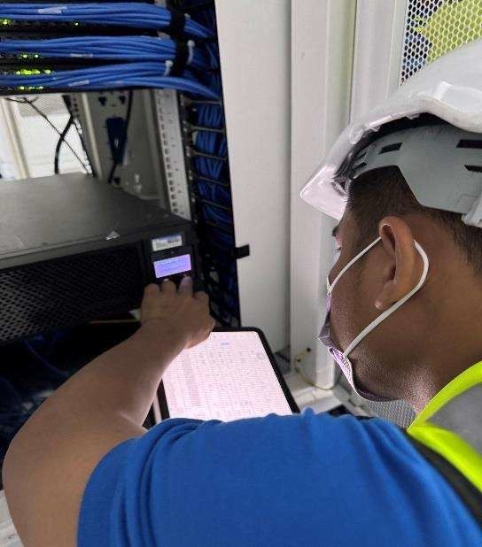 Technician in a hard hat and mask adjusts equipment in a server room with blue network cables.