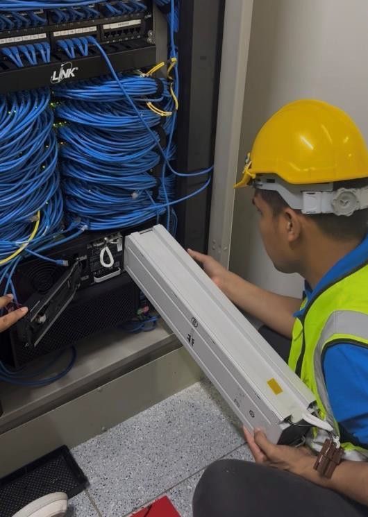 Technician in yellow hard hat inspecting network cabinet with blue cables and a handheld device