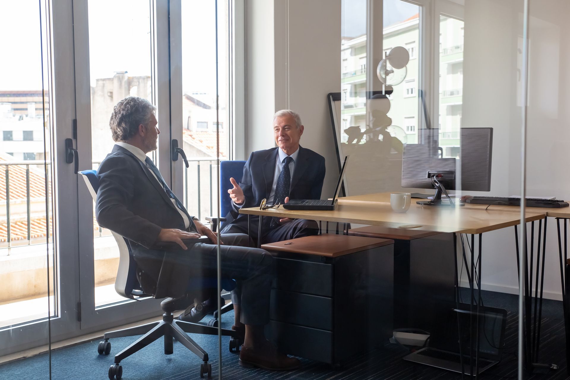 Two men in suits, inside an office with large windows, talking. One gestures with hands.