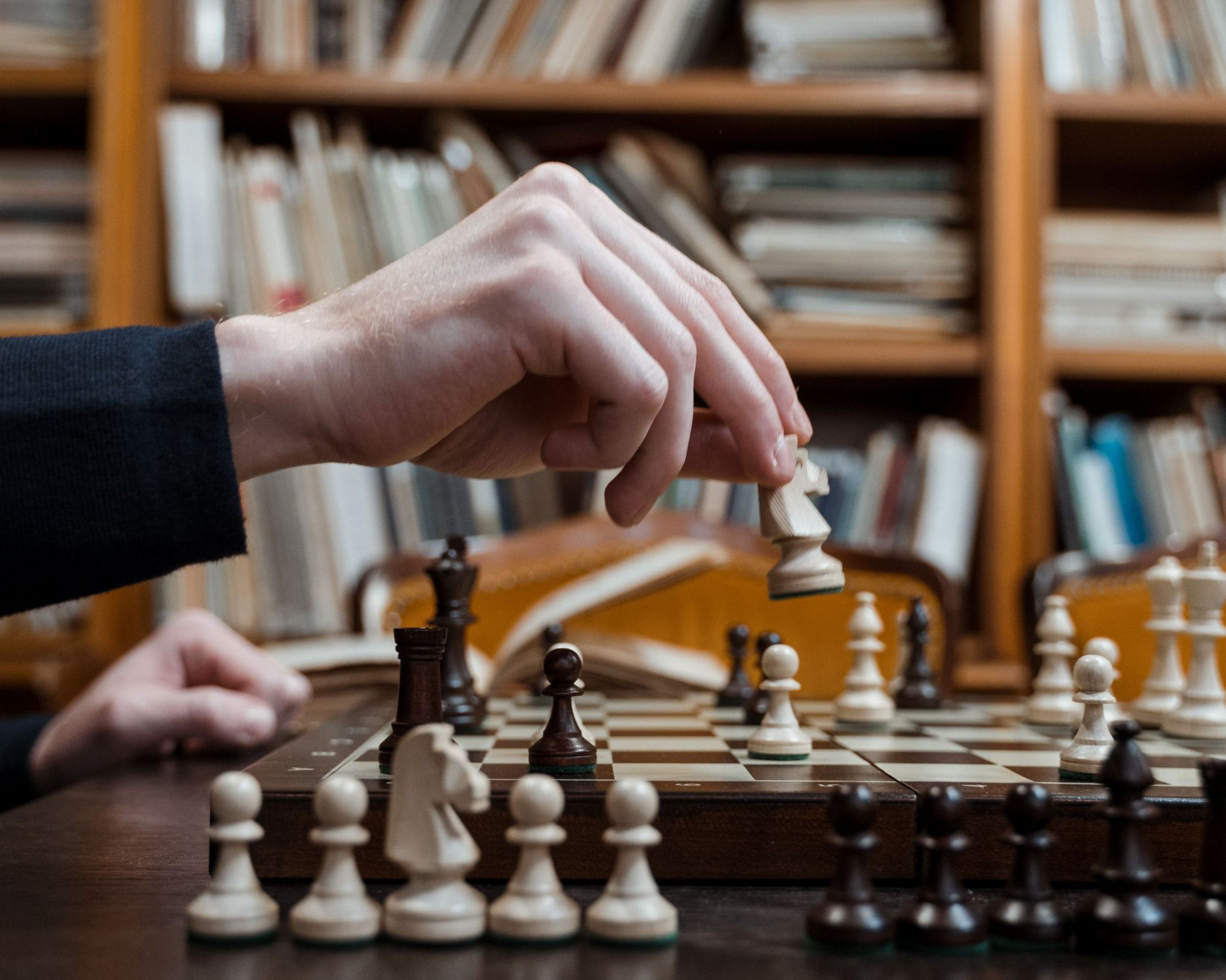 a person holding a white chess knight over a wooden chessboard in a library setting。