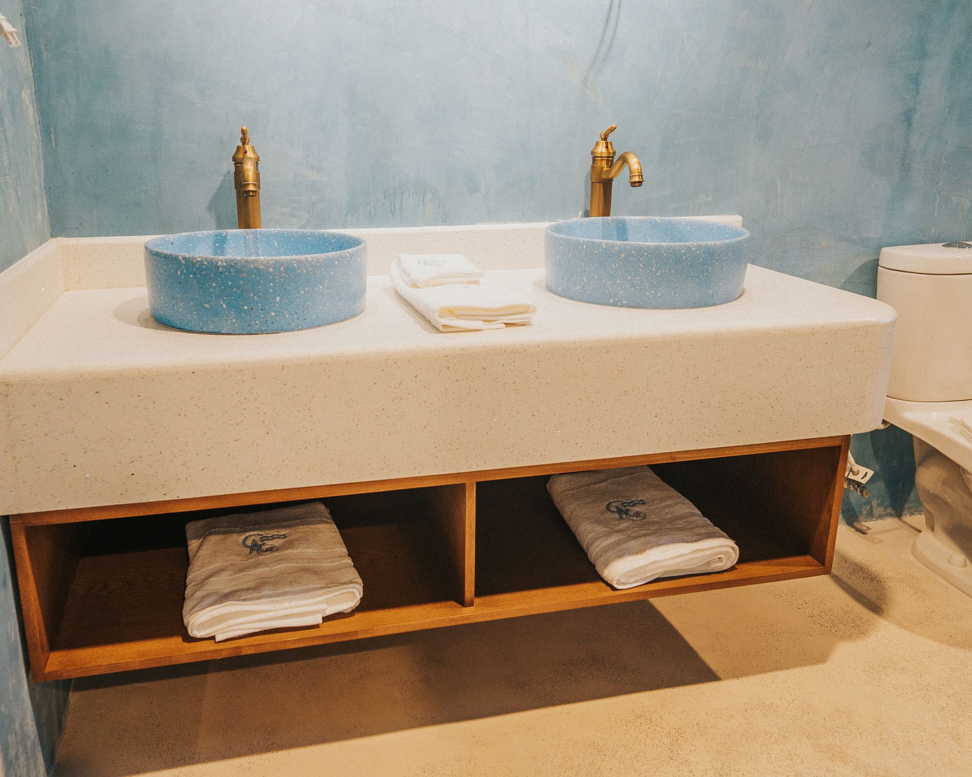 Baño con dos lavabos azules, grifos dorados y un estante de madera con toallas, situado contra una pared azul.