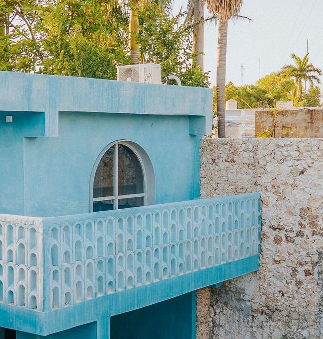 Balcón y fachada de color turquesa con ventana arqueada, fondo de pared de piedra, vegetación tropical.