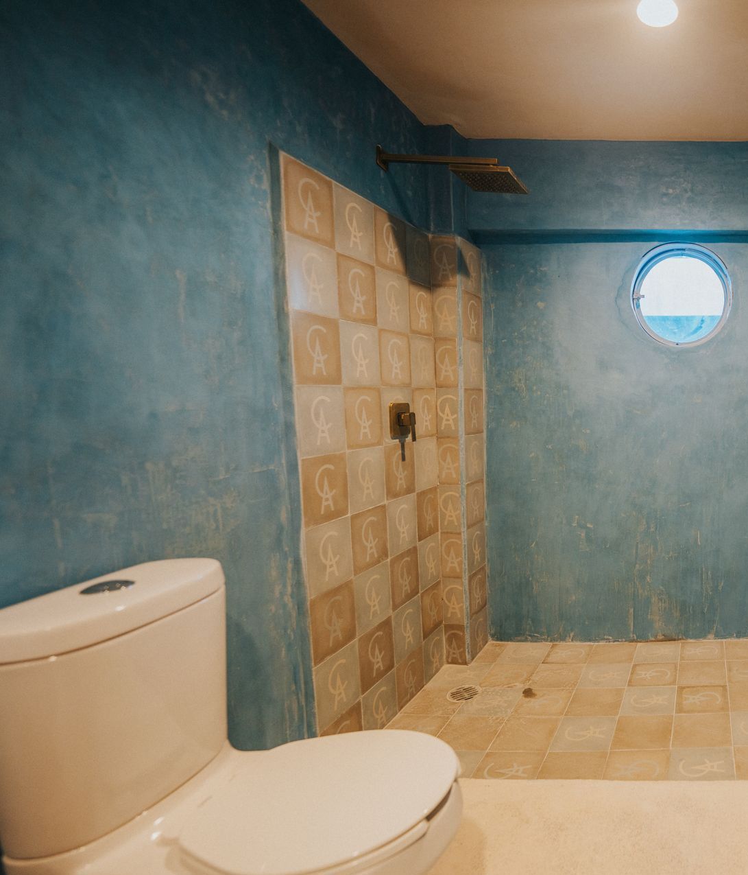 Baño con paredes azules, ducha alicatada, inodoro y ventana redonda.