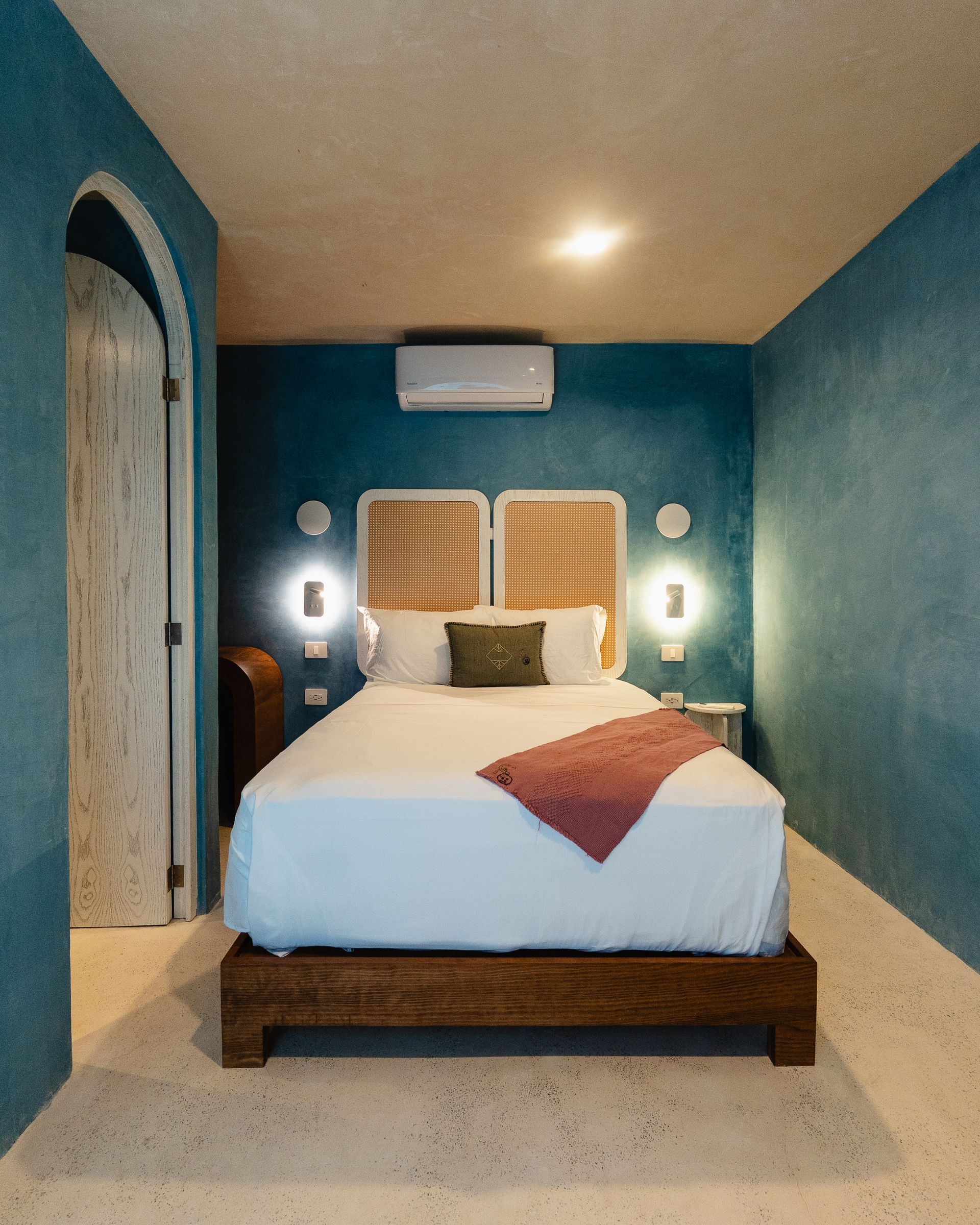Dormitorio con paredes color verde azulado, estructura de cama de madera, cabecero de ratán y colcha blanca con manta rosa.