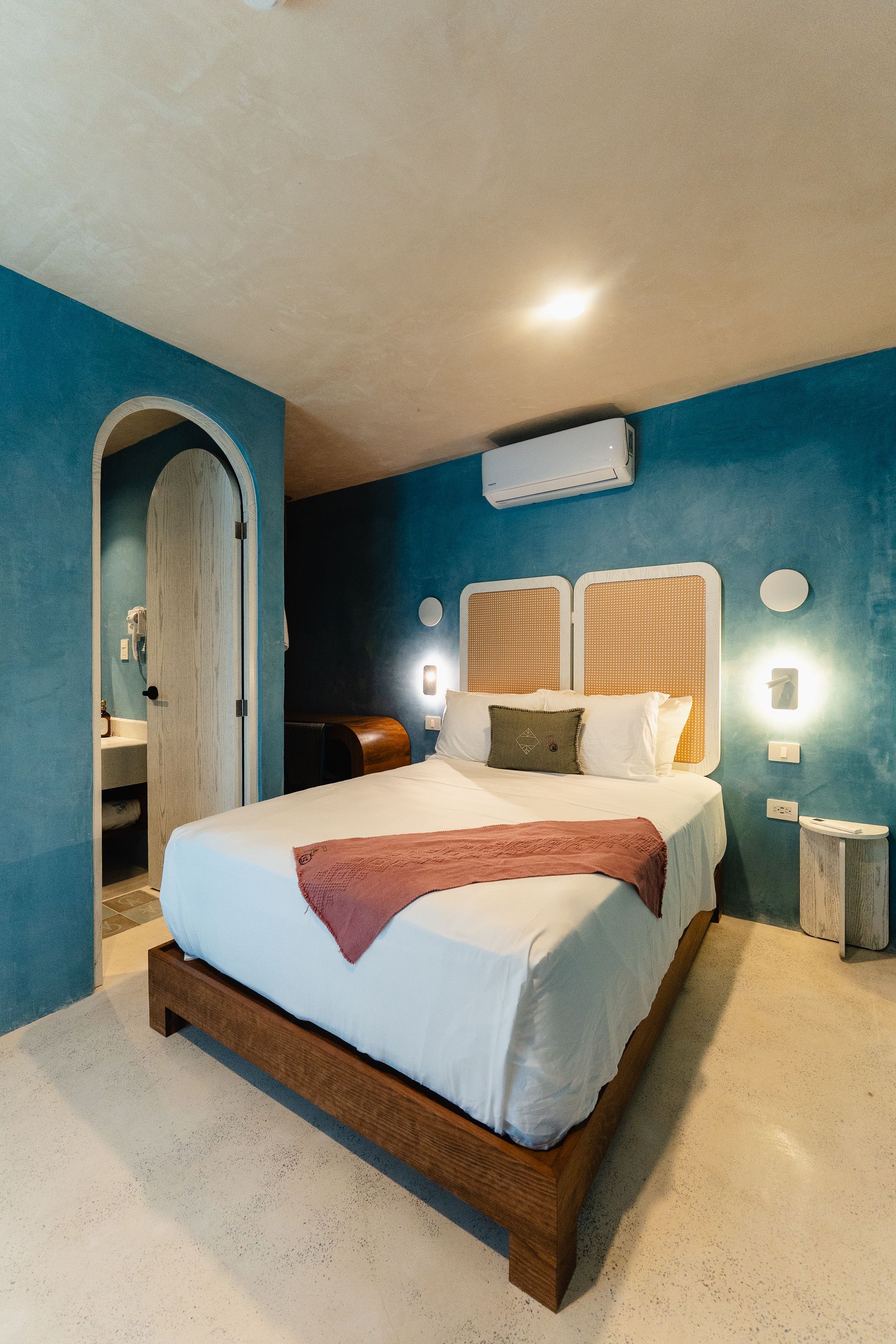 Dormitorio con paredes texturizadas azules, estructura de cama de madera, cabecero tejido y puerta arqueada que da a un baño.