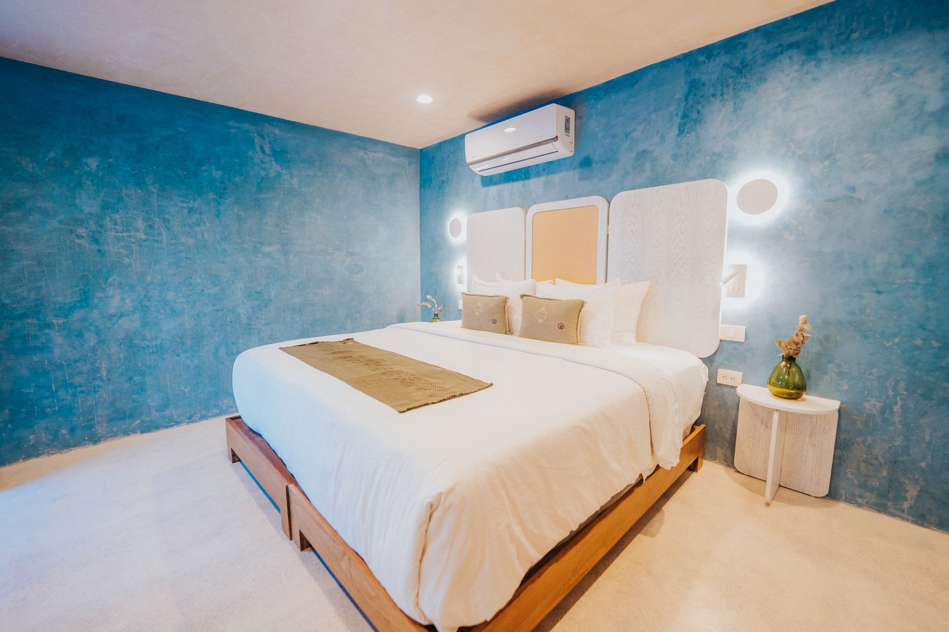 Dormitorio con paredes texturizadas azules, ropa de cama blanca, estructura de cama de madera, luces redondas iluminadas en el cabecero y mesita de noche.