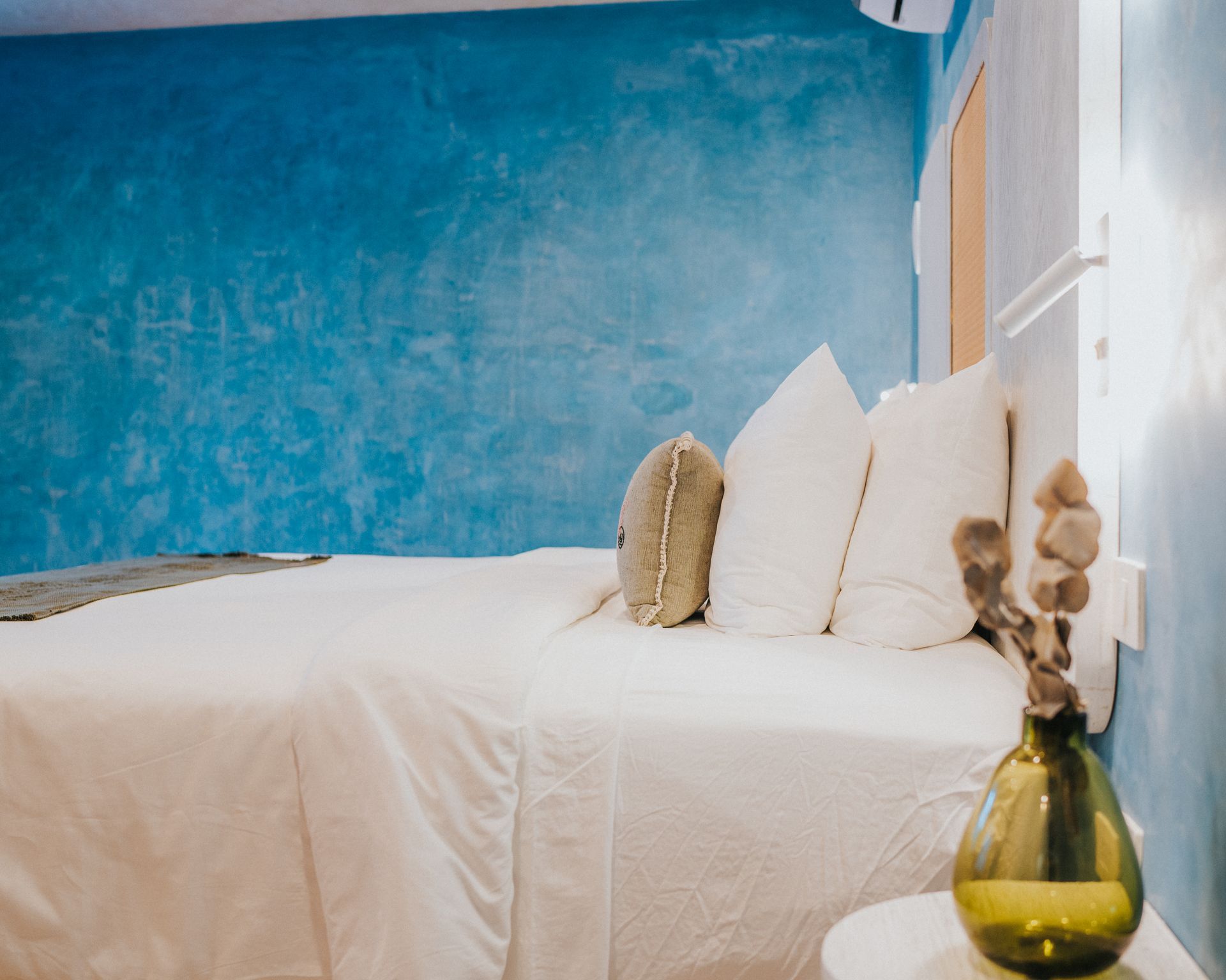 Dormitorio con pared azul texturizada, ropa de cama blanca, cojines decorativos y jarrón.