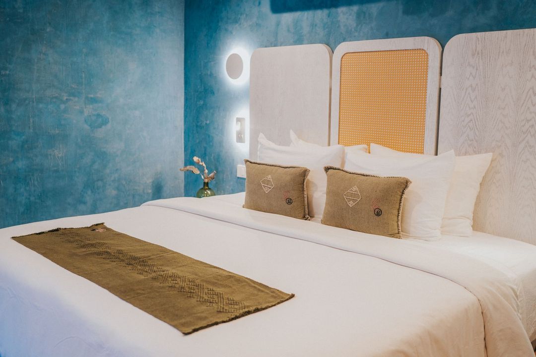 Dormitorio con cama blanca, cojines decorativos, alfombra verde oliva y pared azul texturizada.