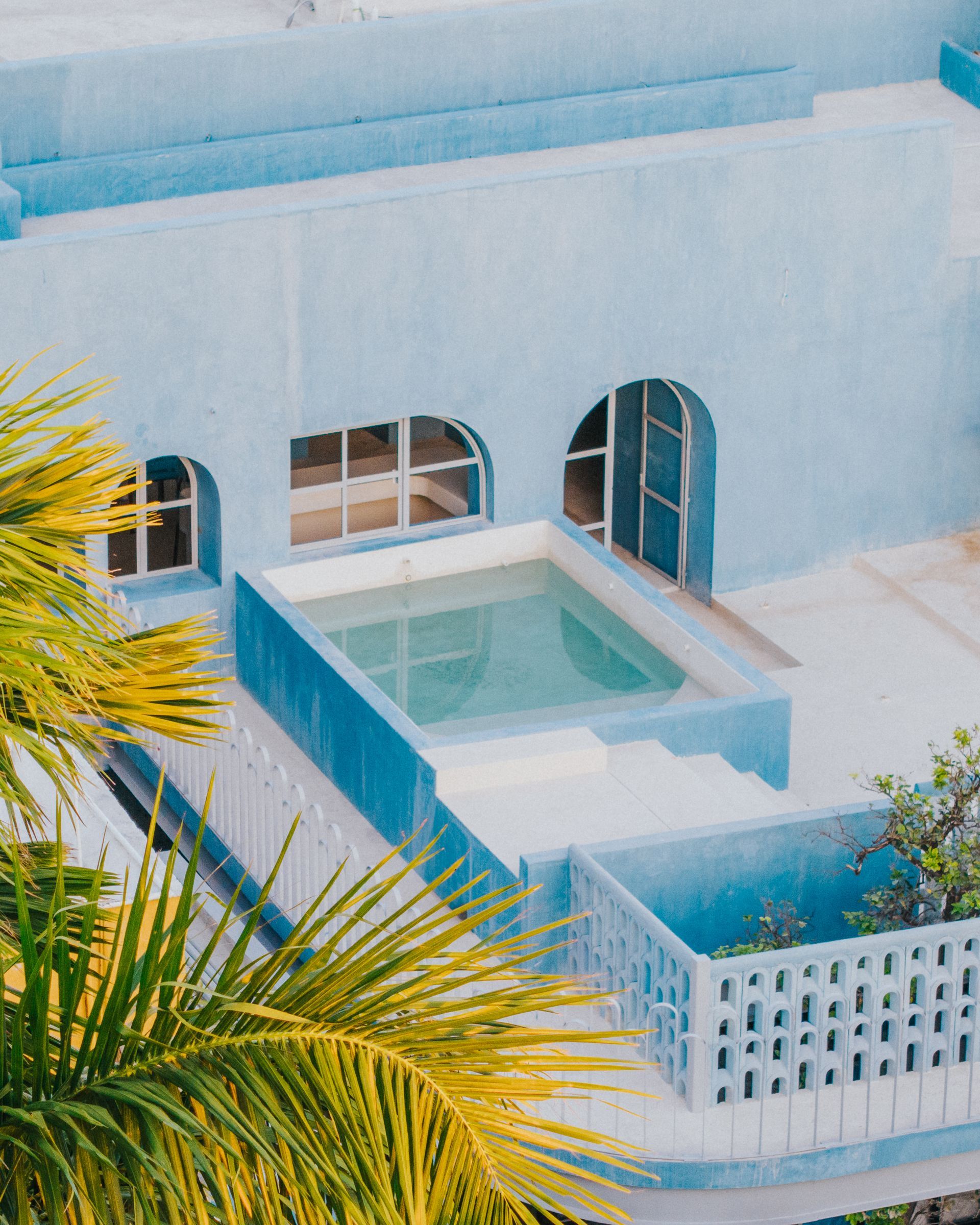 Azotea azul con pequeña piscina y ventanas arqueadas, hojas de palmera en primer plano.