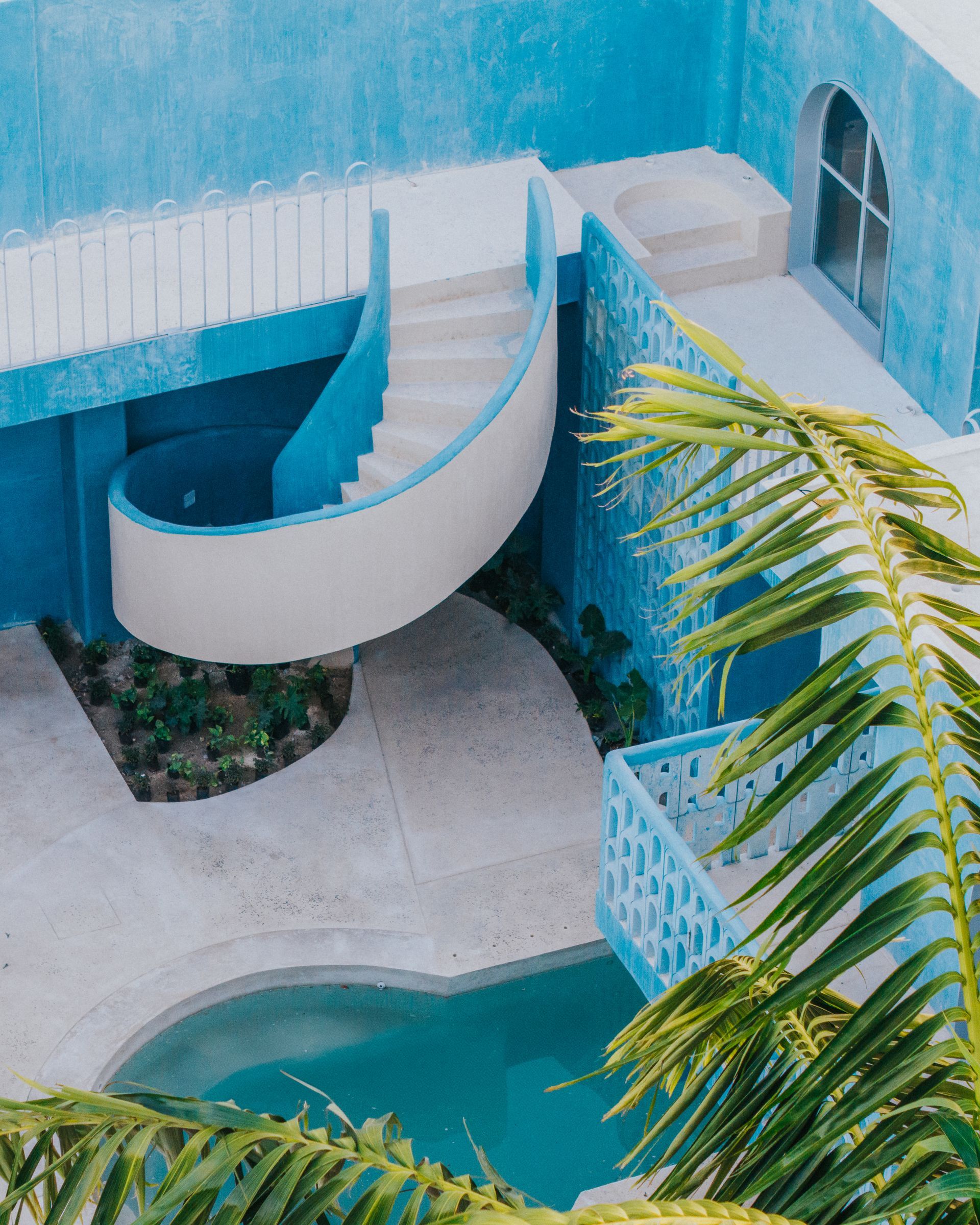 Exterior de un edificio azul con una escalera curva, una pequeña piscina y hojas de palma.