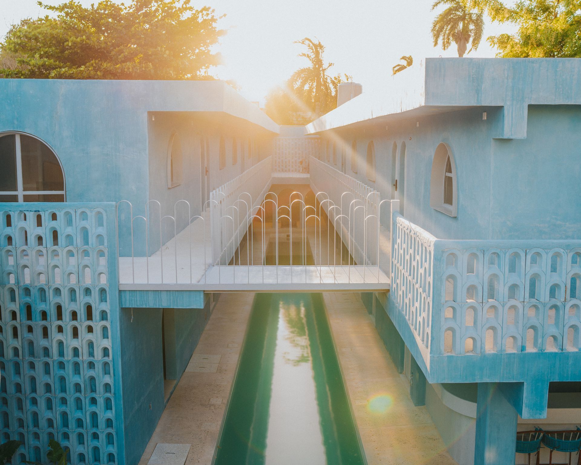 Edificio azul de estilo moderno de mediados de siglo con puente sobre una larga piscina; destellos de luz solar brillante.