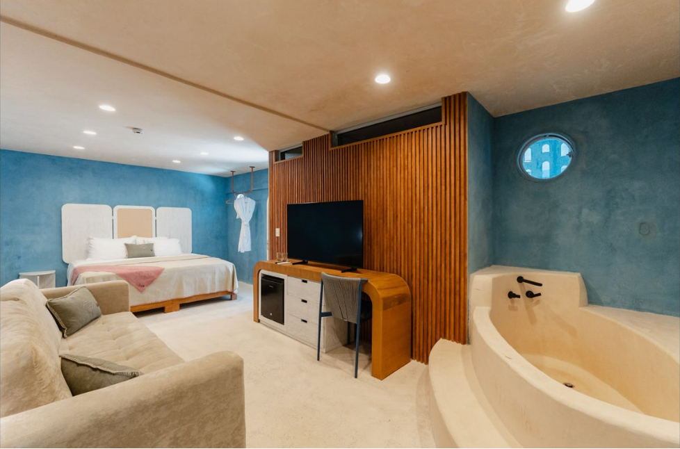 Habitación de hotel con cama, TV, sofá, escritorio y bañera; paredes azules, detalles de madera.
