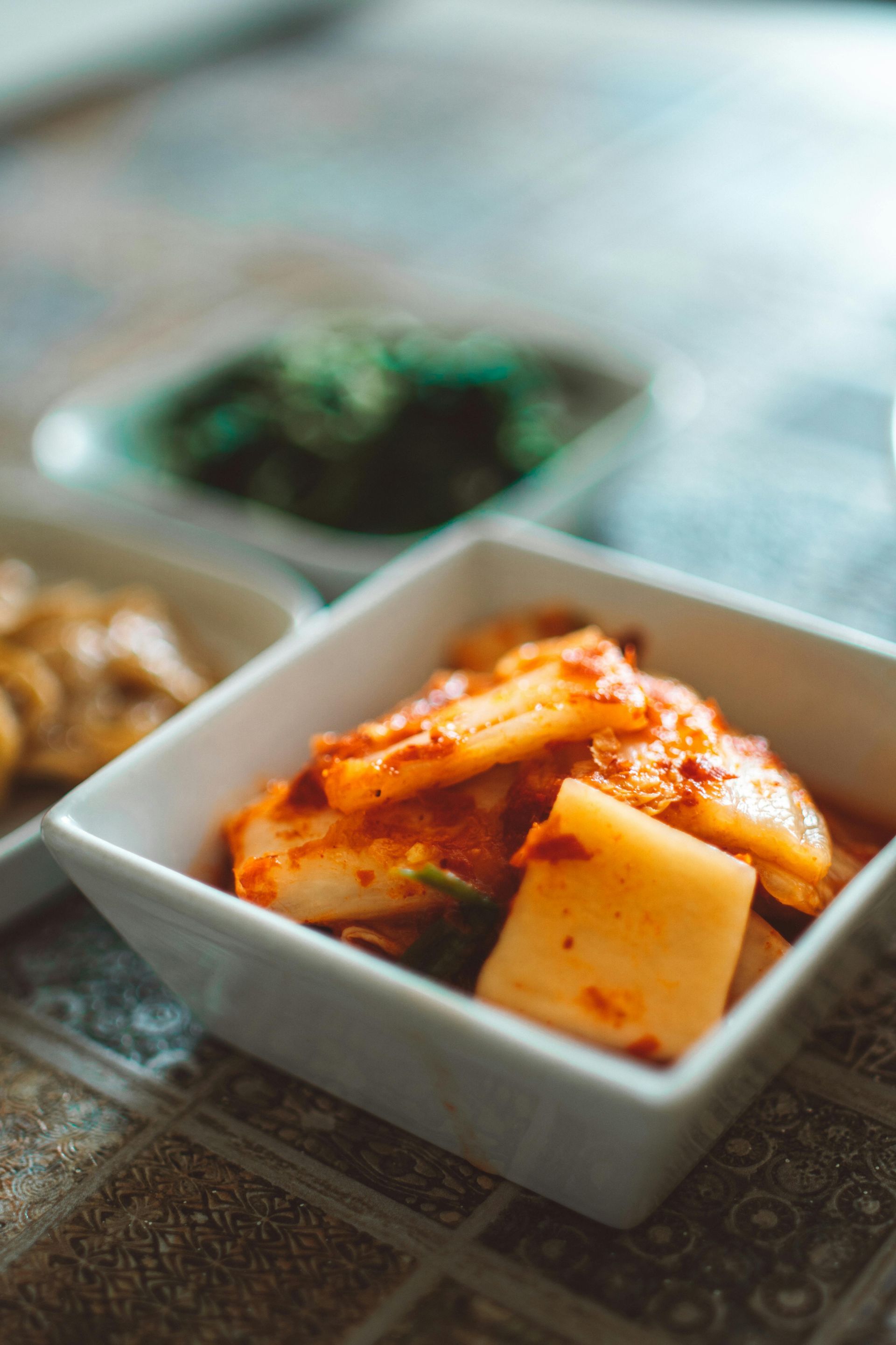 Kimchi en un bol cuadrado blanco sobre una mesa estampada; otros platos pequeños aparecen borrosos al fondo.