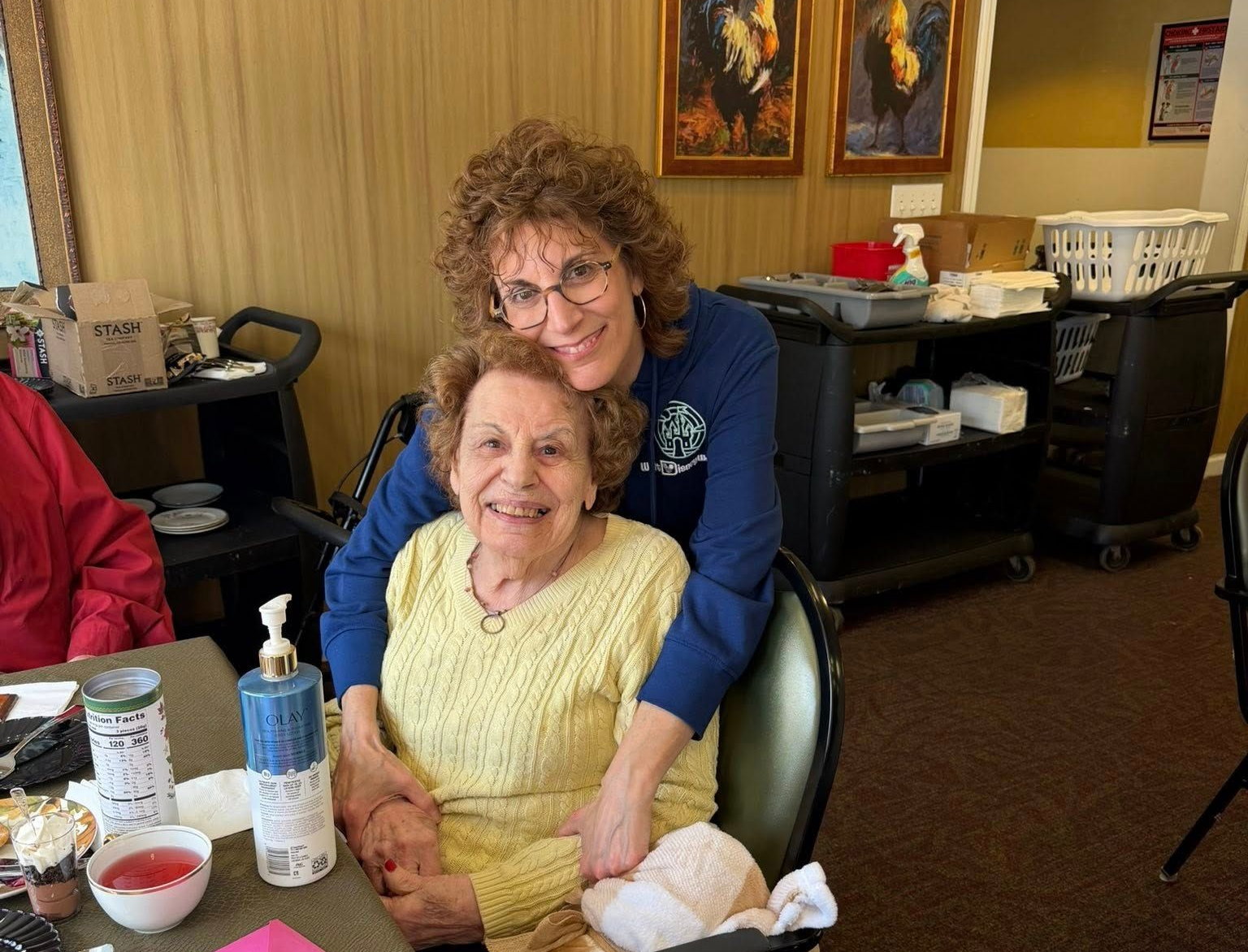 Two people smiling at a table in a cozy room with framed art and office furniture.