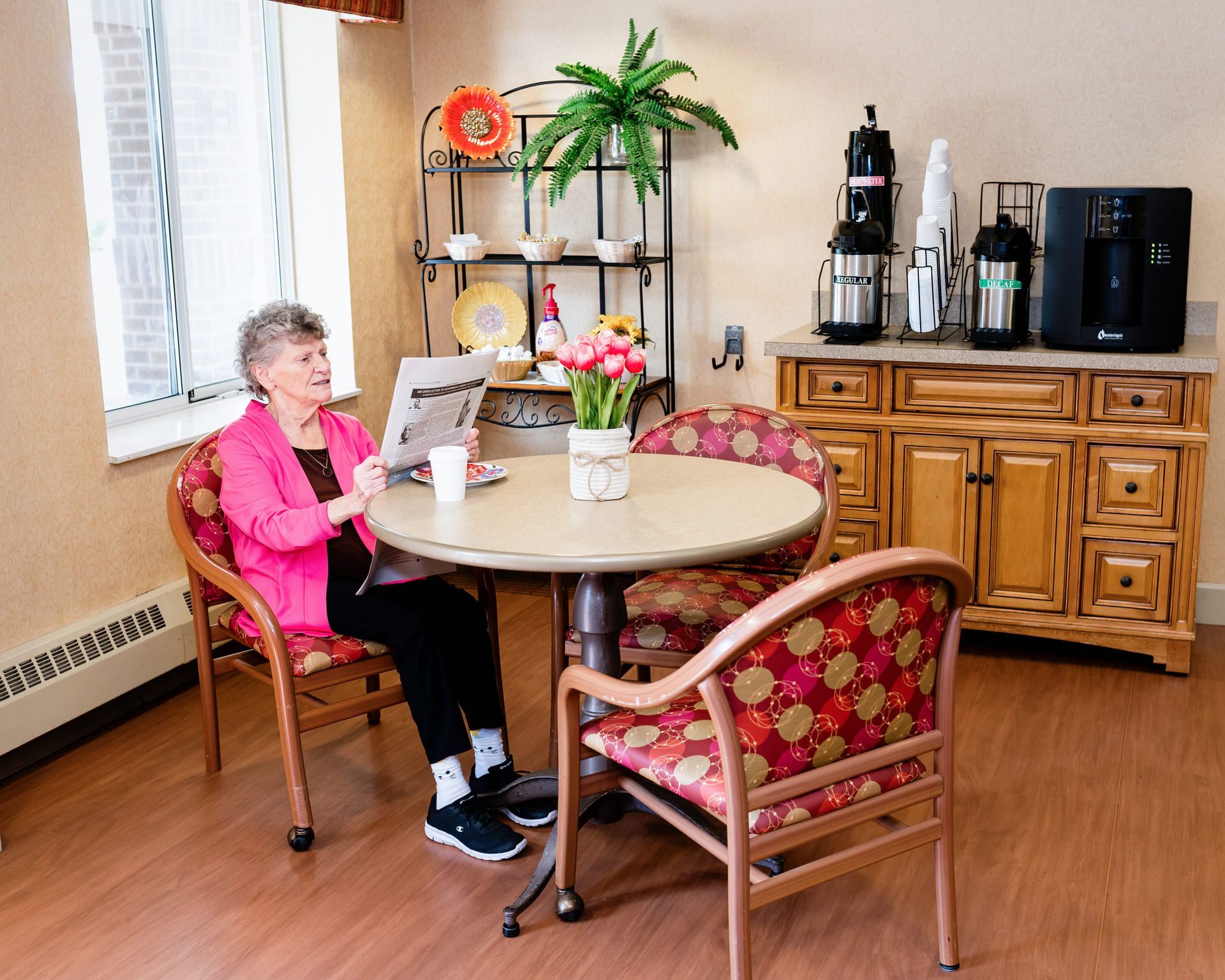 A person in a bright pink blazer sits at a circular table, reading a newspaper in a bright, modern dining area.