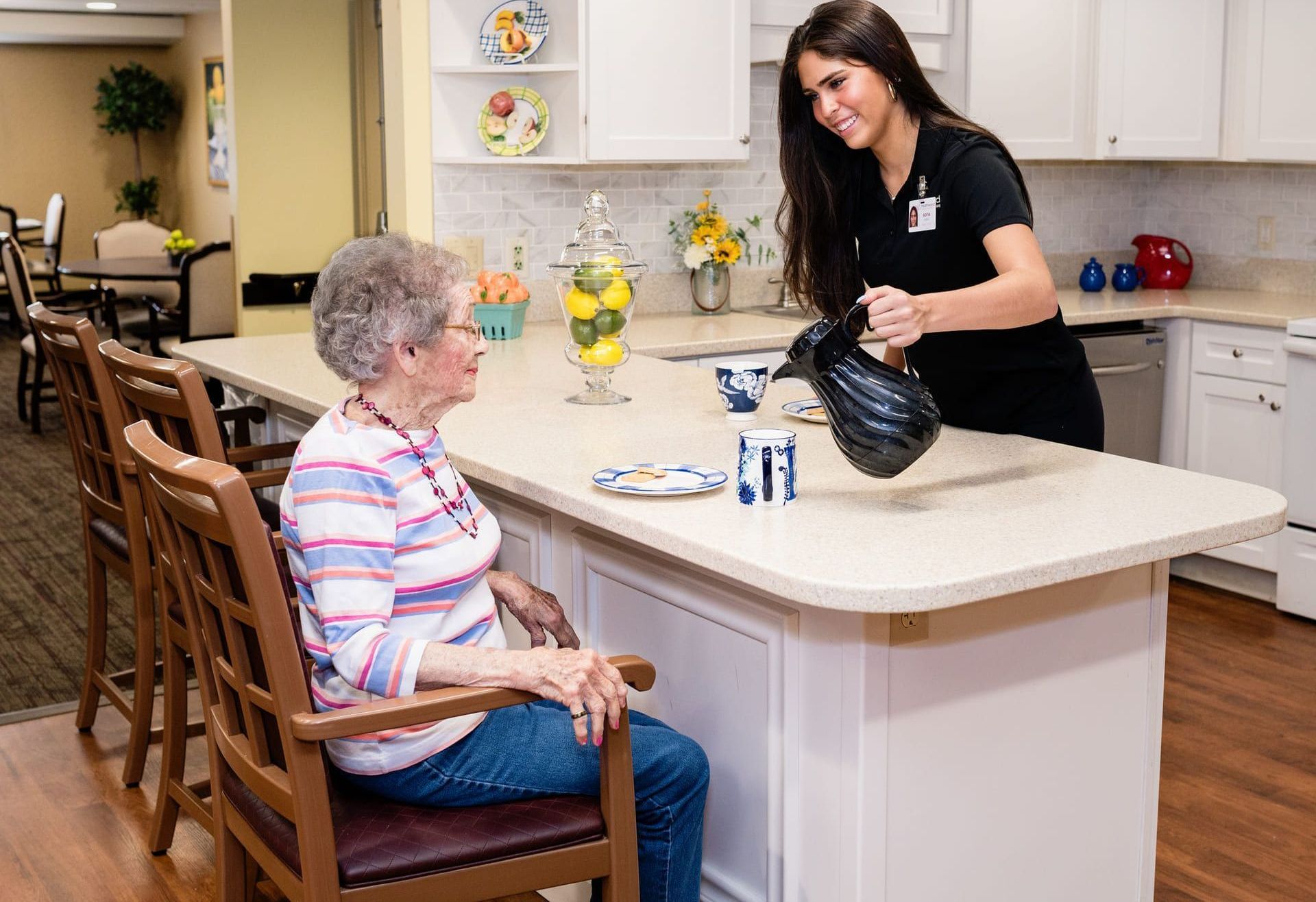 Caregiver pours water for an elderly woman seated at a kitchen island.