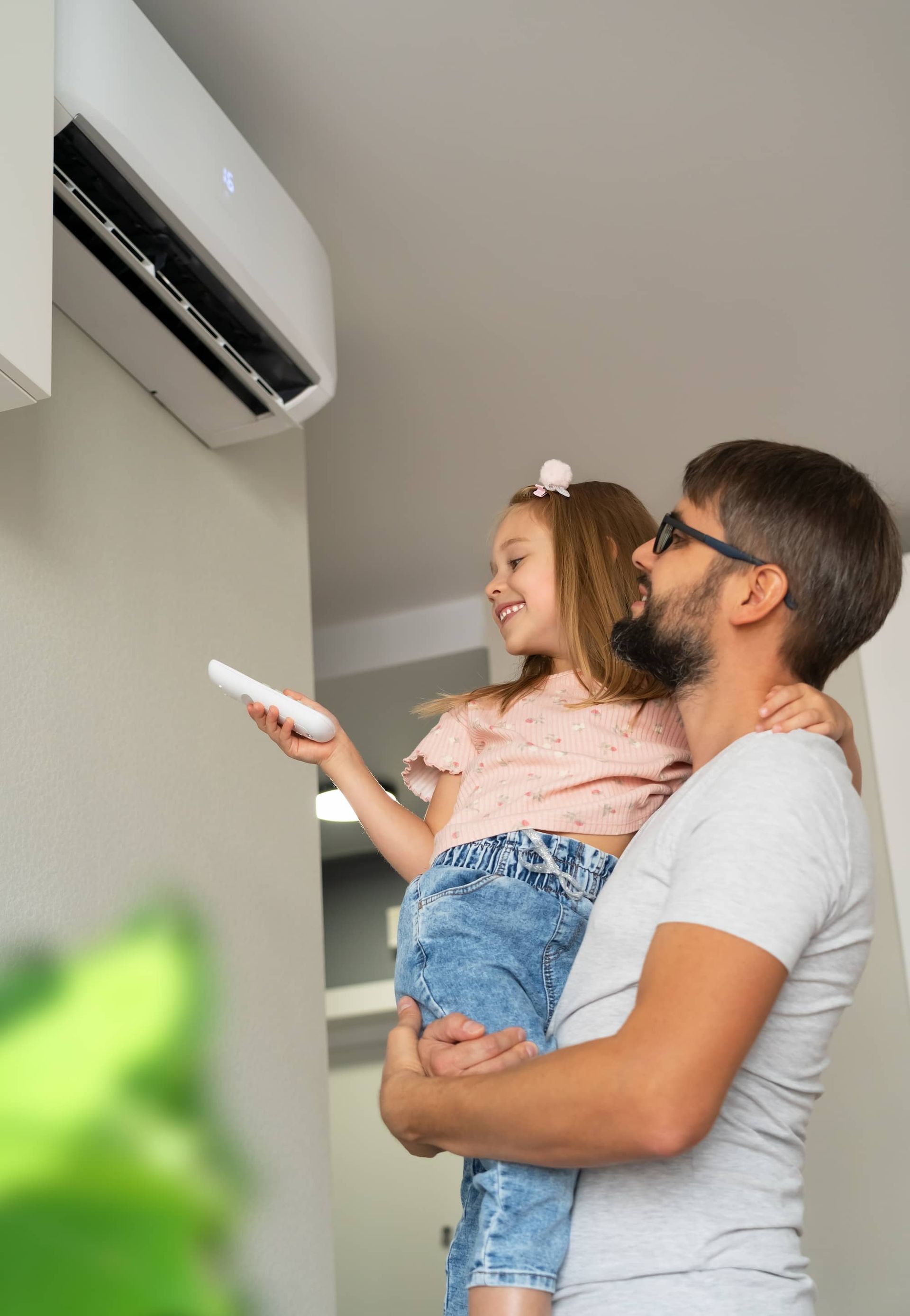 A Man is Holding a Little Girl in His Arms While She Holds a Remote Control in Front of an Air Conditioner — Elements Air & Refrigeration In Westcourt, QLD