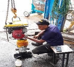 Un trabajador repara una máquina roja al aire libre, junto a mangueras y herramientas, en una obra húmeda.