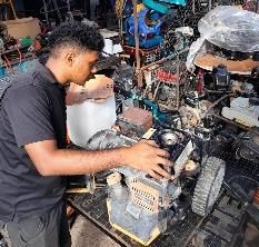Una persona repara un motor pequeño en un taller desordenado, rodeada de herramientas y maquinaria.