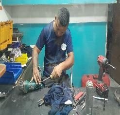 Mecánico trabajando en una herramienta eléctrica verde en un banco de trabajo desordenado con herramientas y un taladro rojo cerca.