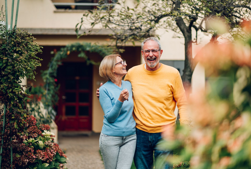 Smiling couple walking together in a garden path, with a house and flowers in the background.