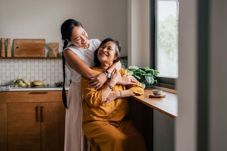 Two people embracing in a bright kitchen, smiling by a window with houseplants on the counter