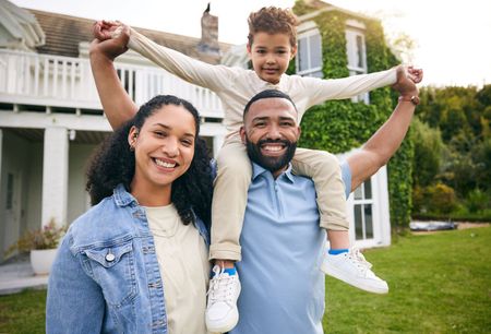 Smiling family of three outdoors, with a child on a man's shoulders in a yard by a house
