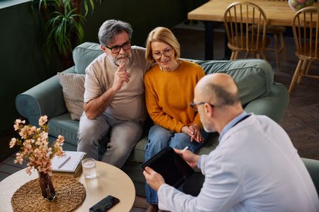 Three people talking on a couch in a cozy living room, with a coffee table and flowers in the foreground.