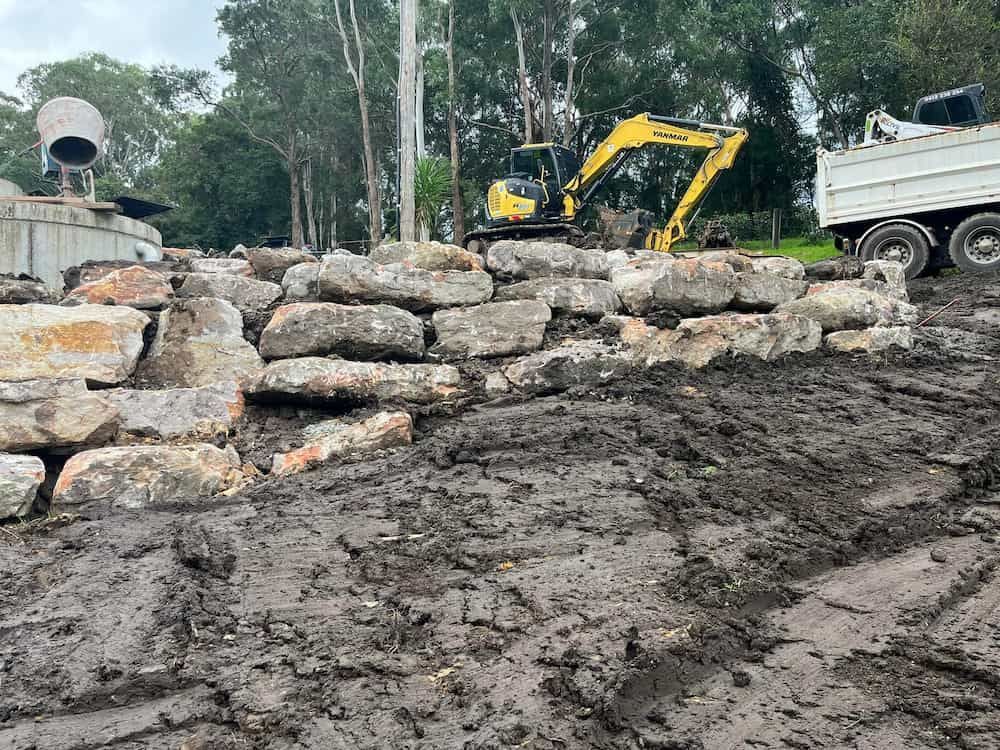 A Yellow Excavator is Working on a Pile of Rocks Next to a Dump Truck — BeCivil in Tomerong, NSW