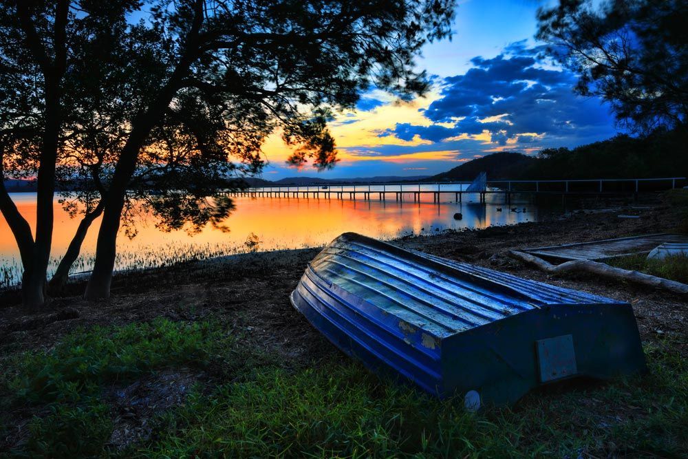 A Boat Is Sitting On The Shore Of A Lake At Sunset — Coast 2 Coast Batteries in Kincumber, NSW