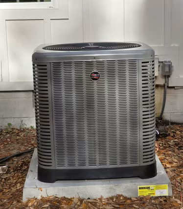 Outdoor air conditioning unit beside a house wall on a concrete pad, with leaves around it