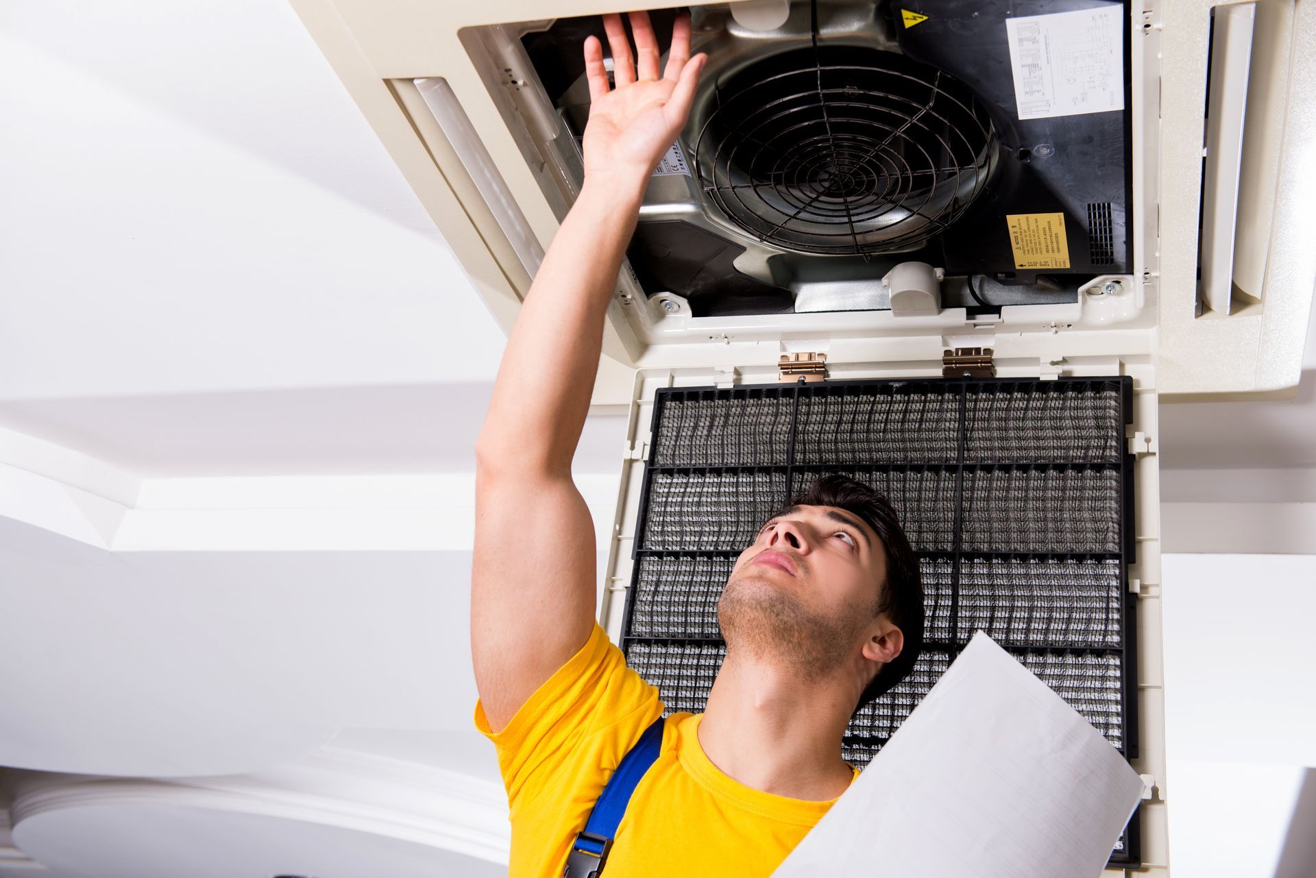 Technician in yellow shirt inspecting a ceiling air conditioning unit with a clipboard.