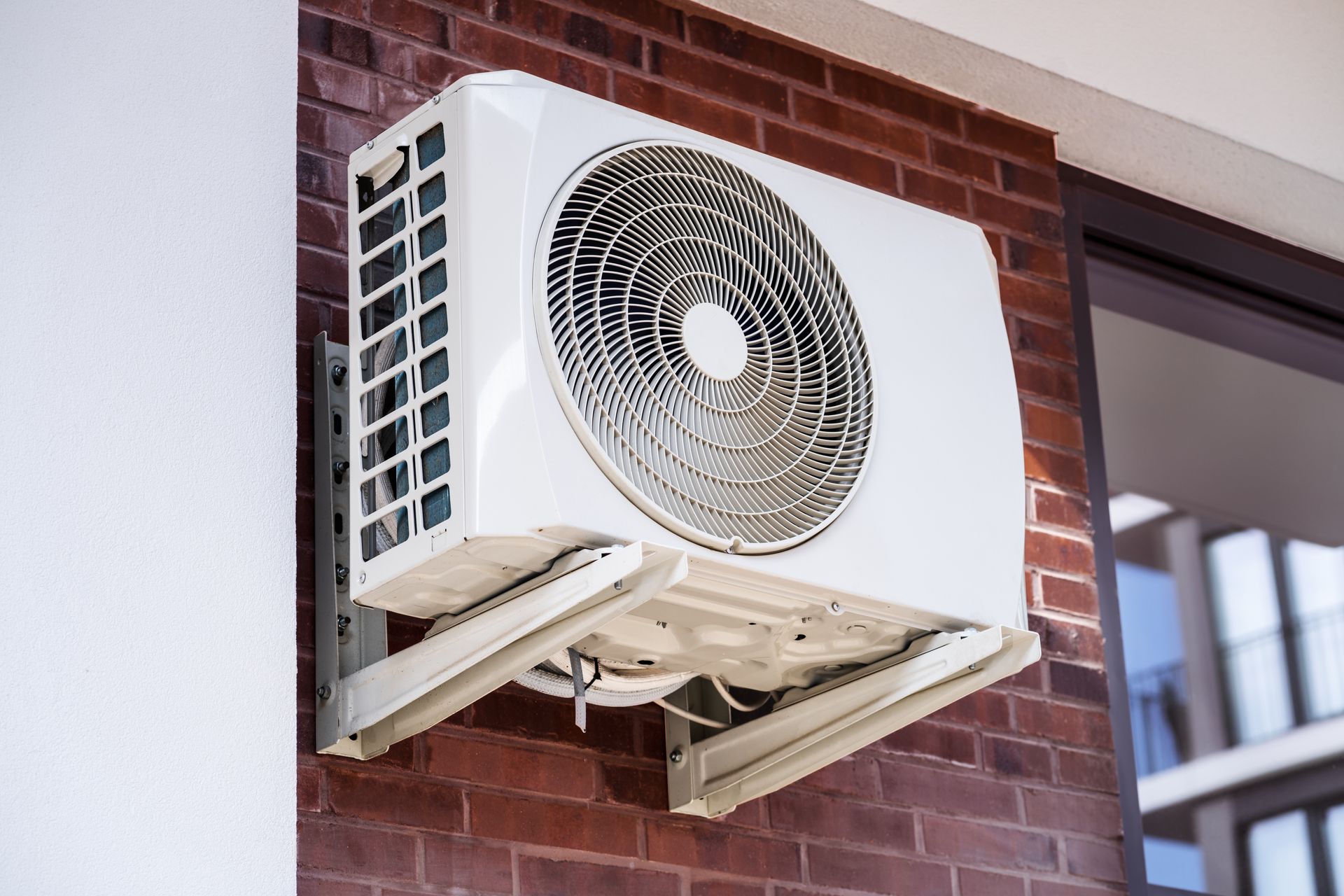Outdoor air conditioner unit mounted on a brick wall, with a window beside it