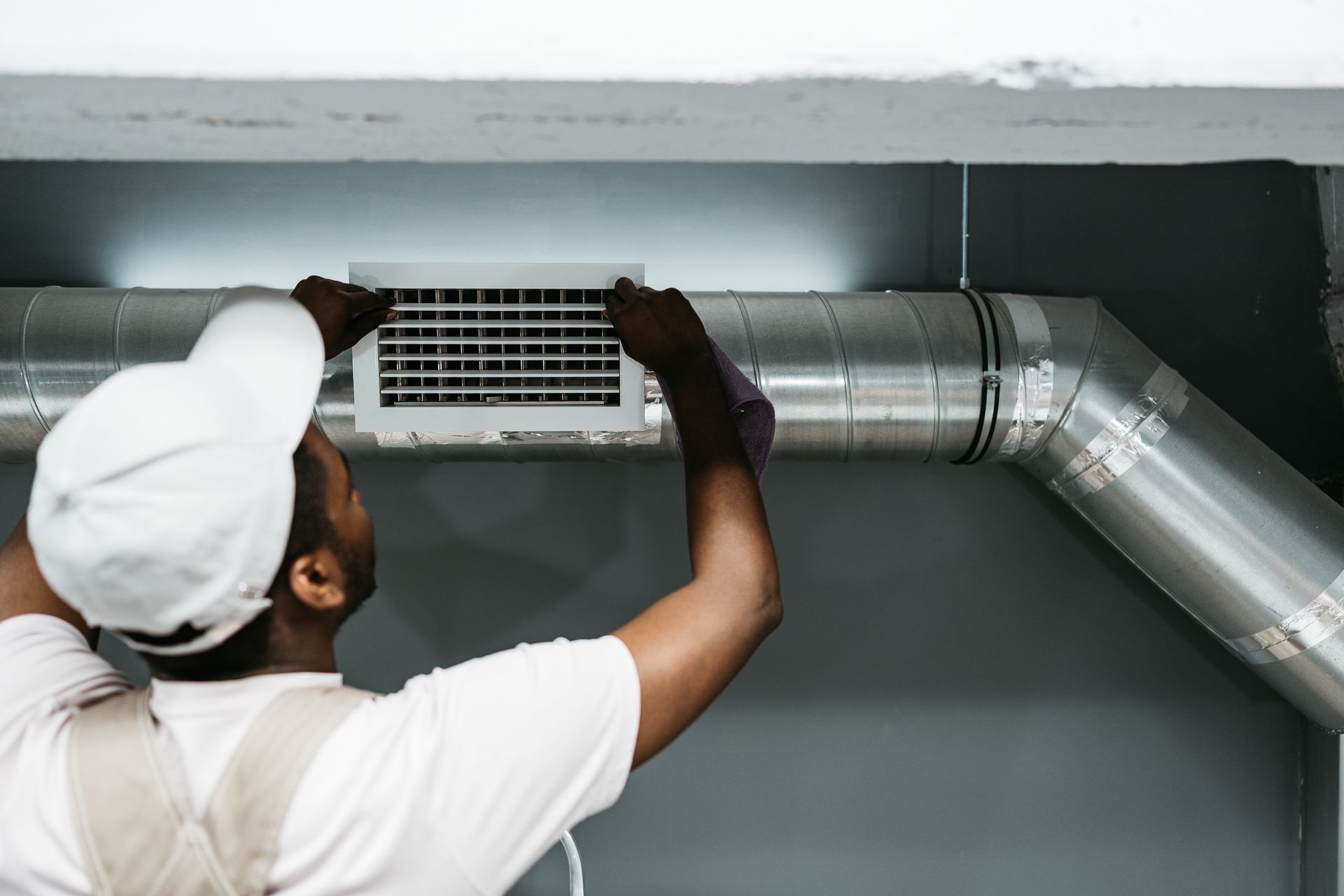 Worker cleaning a ceiling vent beside metal ductwork in a gray room
